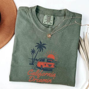 May include: Olive green t-shirt with a graphic of a red van, palm trees, and a setting sun. The text "California Dreamin'" is printed below the graphic. A gold necklace and a brown hat are also visible.