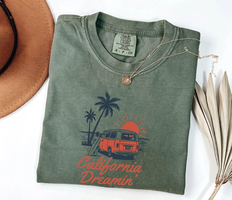 May include: Olive green t-shirt with a graphic of a red van, palm trees, and a setting sun. The text "California Dreamin'" is printed below the graphic. A gold necklace and a brown hat are also visible.