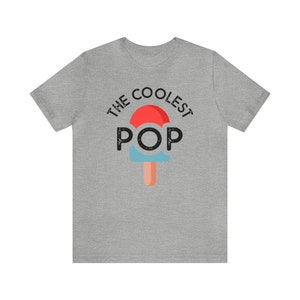 Puede incluir: Camiseta gris jaspeado con la frase "THE COOLEST POP" sobre un gr&aacute;fico de paleta helada. La paleta tiene la parte superior roja, el centro azul y un palo naranja claro. La camiseta es de cuello redondo y manga corta.