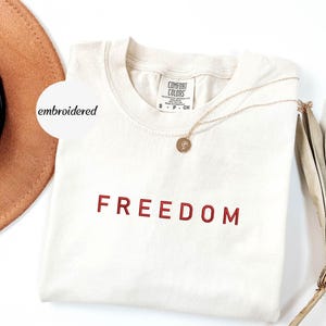 May include: Cream-coloured t-shirt with the word "FREEDOM" embroidered in red. A gold necklace with a pendant rests on the shirt. A brown hat and dried plant are in the background. The shirt has a tag that says "Comfort Colors".