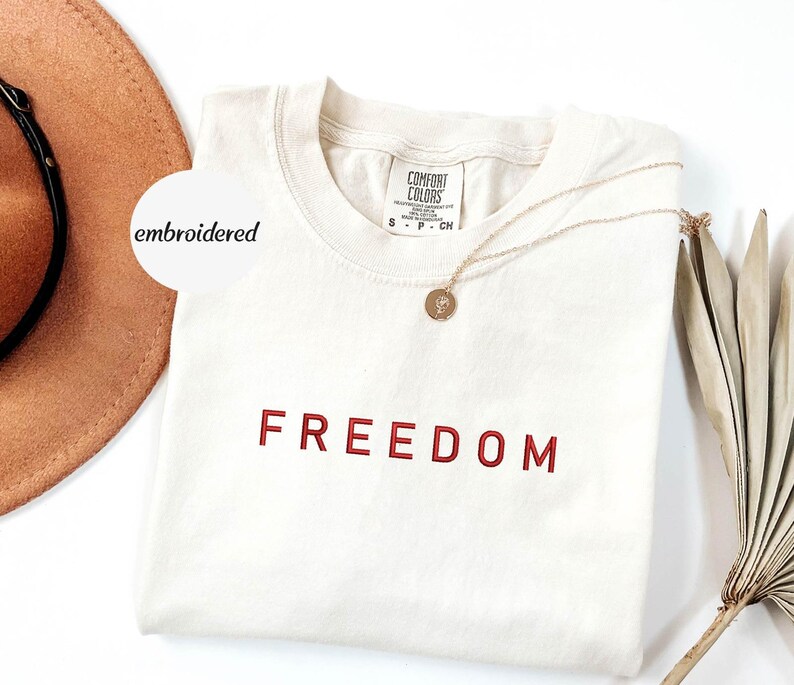 May include: Cream-coloured t-shirt with the word "FREEDOM" embroidered in red. A gold necklace with a pendant rests on the shirt. A brown hat and dried plant are in the background. The shirt has a tag that says "Comfort Colors".