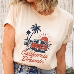 May include: Cream-colored t-shirt featuring a retro graphic of a red and white van parked on a beach with palm trees and a setting sun. The text "California Dreamin'" is in red script.