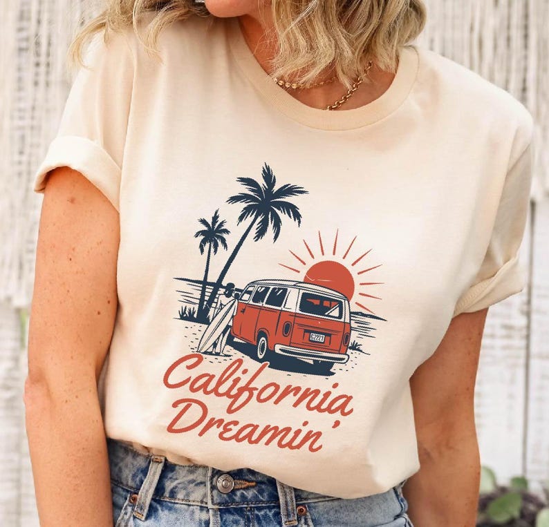 May include: Cream-colored t-shirt featuring a retro graphic of a red and white van parked on a beach with palm trees and a setting sun. The text "California Dreamin'" is in red script.