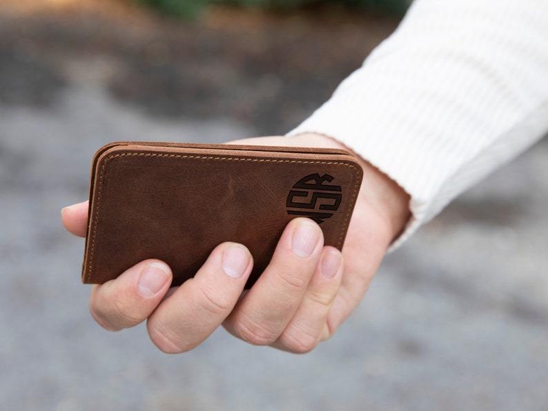 May include: A brown leather wallet with a monogrammed circle on the front. The wallet is being held in a hand.