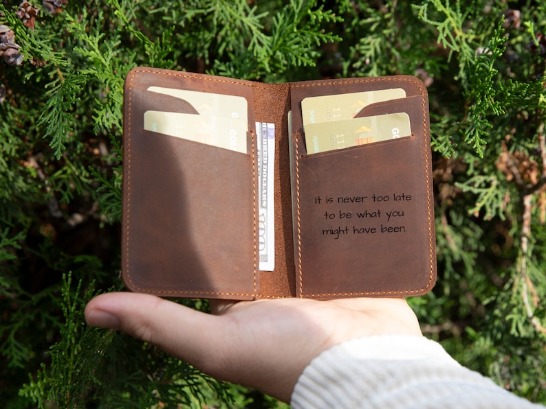 May include: A brown leather wallet with a quote embossed on the front: "It is never too late to be what you might have been". The wallet is open and has several cards and cash inside.