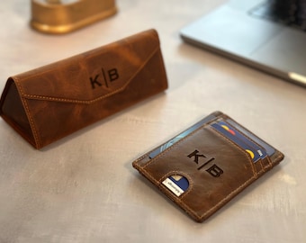 May include: A brown leather glasses case and a matching wallet, both with the initials "K|B" engraved. The wallet holds several cards. The items are on a light gray surface, with a laptop visible in the background.