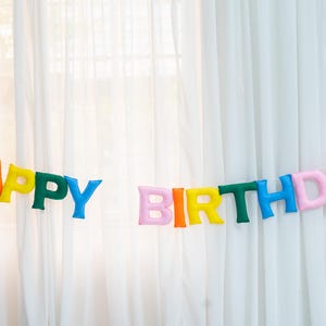 Può includere: Uno striscione di feltro colorato con le parole "Happy Birthday" infilate su uno sfondo bianco.