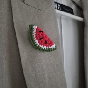 Könnte beinhalten: Ein gehäkeltes Wassermelonen-Scheiben-Anstecknadel in Rot, Weiß, Grün und Schwarz, befestigt an einem grauen Blazer. Der Blazer ist aus einem strukturierten Stoff gefertigt und hängt an einem weißen Kleiderbügel. Das Accessoire ist handgefertigt.