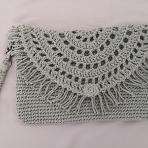 Könnte beinhalten: Eine hellgraue gehäkelte Clutch mit Fransen und einem gehäkelten Blumenakzent. Die Tasche hat einen langen Riemen.