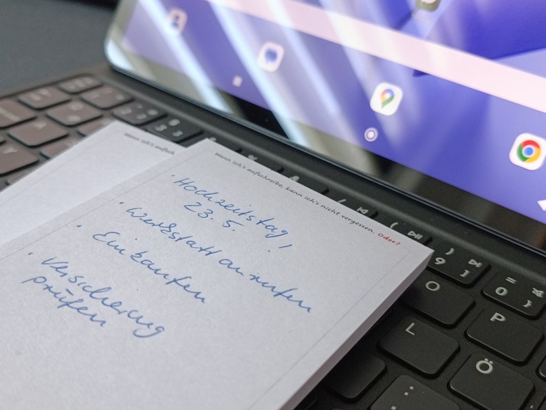 May include: A white notepad with blue handwritten text on it. The text says "Hochzeitstag! 23.5. Werkstatt anrufen. Ein kaufen. Versicherung pr&uuml;fen."