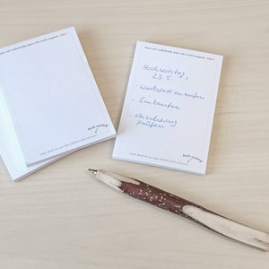 May include: A stack of white notepad paper with a handwritten to-do list on the top sheet. The list includes "Hochzeitstag 23.5." and "Werkstatt anrufen". The notepad has the text "So what, that I am the last letter, I will not apologize!" and "Auch wichtig!" printed on the bottom. A brown and white wooden pen is lying on a light wood surface.