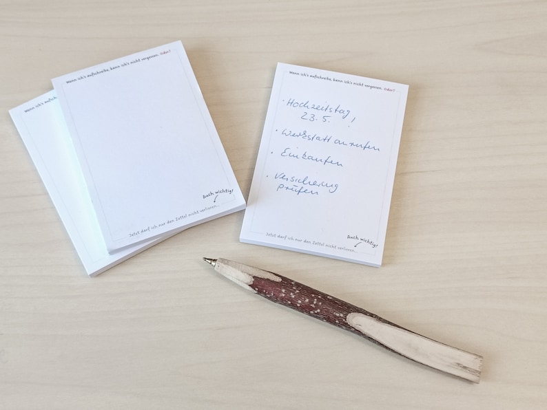 May include: A stack of white notepad paper with a handwritten to-do list on the top sheet. The list includes "Hochzeitstag 23.5." and "Werkstatt anrufen". The notepad has the text "So what, that I am the last letter, I will not apologize!" and "Auch wichtig!" printed on the bottom. A brown and white wooden pen is lying on a light wood surface.