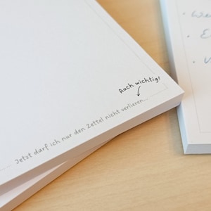 May include: A white notepad with the text "Auch wichtig!" and "Jetzt darf ich nur den Zettel nicht verlieren..." written on it. The notepad is stacked on top of another notepad.