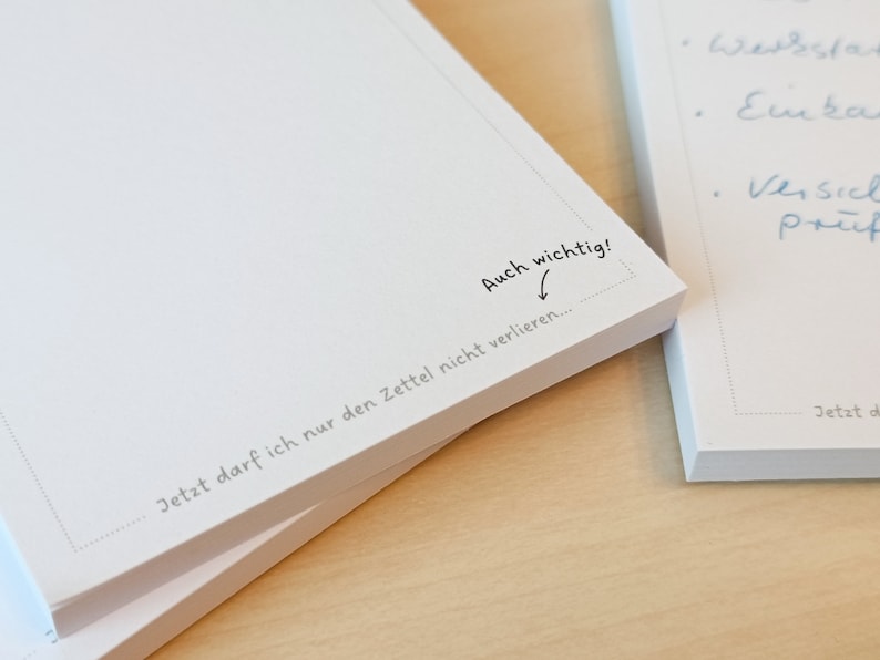 May include: A white notepad with the text "Auch wichtig!" and "Jetzt darf ich nur den Zettel nicht verlieren..." written on it. The notepad is stacked on top of another notepad.