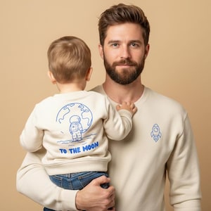 Könnte beinhalten: Ein Mann hält ein Kind, beide tragen passende cremefarbene Sweatshirts. Das Sweatshirt des Kindes zeigt eine blaue Astronauten-Grafik mit dem Text "TO THE MOON". Das Sweatshirt des Mannes hat eine blaue Raketen-Grafik. Neutraler Hintergrund.