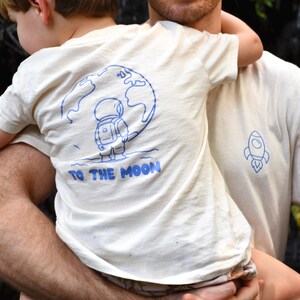 Könnte beinhalten: Off-white T-Shirts mit blauen Grafiken. Eines zeigt eine Rakete, das andere einen Astronauten auf dem Mond mit dem Text "TO THE MOON". Die Shirts werden von einem Kind und einem Erwachsenen getragen.
