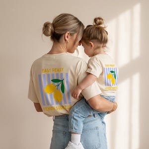 Könnte beinhalten: Cremefarbene T-Shirts mit einem Zitronen-Grafik und dem Text "EASY PEASY LEMON SQUEEZY". Die Shirts haben einen blau gestreiften Hintergrund. Das Bild zeigt eine Mutter und ein Kind, die die Shirts tragen.