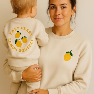 Könnte beinhalten: Eine Frau hält ein Kind, das passende cremefarbene Sweatshirts trägt. Das Sweatshirt des Kindes hat eine Zitronengrafik mit dem Text "Easy Peasy Lemon Squeezy". Das Sweatshirt der Frau hat eine einzelne Zitronengrafik.