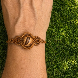 Op de afbeelding: Een bruine macramé armband met een centrale ronde kraal met een beige en donkerbruin marmerpatroon. De armband is gemaakt van geknoopt koord, wat zorgt voor een symmetrisch, decoratief ontwerp dat de pols omcirkelt met ingewikkelde lussen.