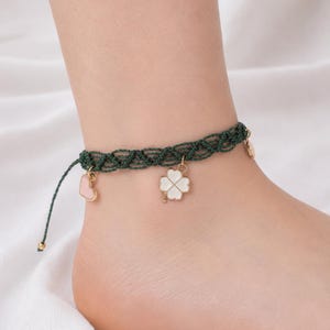 Puede incluir: Una tobillera de macramé verde oscuro con un diseño de cordón trenzado y dijes colgantes. Los dijes incluyen un trébol de cuatro hojas blanco y un pequeño corazón rosa claro. La tobillera tiene extremos de cordón ajustables con cuentas.