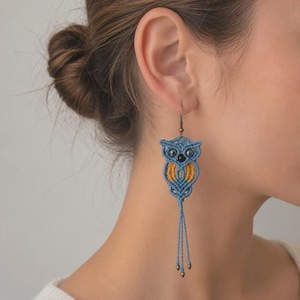 Op de afbeelding: Blauwe en gele macramé uil oorbellen met een bungelend kwastje ontwerp. De oorbellen hebben een gedetailleerde uilvorm met zwarte kraalogen en een gele kern. Ze hangen aan een bronzen haak.