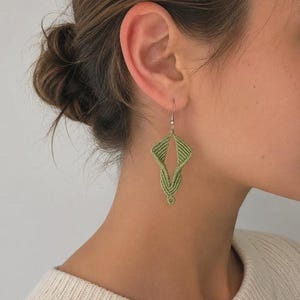May include: A close-up of a person's ear wearing a pair of green macrame earrings. The earrings have a geometric design and are suspended from silver-colored hooks. The person is wearing a white sweater.