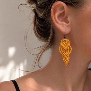 Può includere: Un orecchino in macramè color giallo senape caratterizzato da un intricato design a cordoncino annodato a forma di goccia. L'orecchino pende da un gancio in metallo scuro, mostrando un motivo intrecciato dettagliato che si restringe in punta.