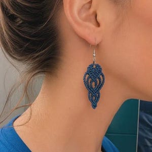 Puede incluir: Un pendiente de macramé azul oscuro con un intrincado diseño de cordón anudado en forma de lágrima. El pendiente cuelga de un gancho de metal plateado. El patrón anudado consta de múltiples bucles y hebras tejidas simétricamente.