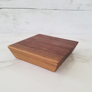 May include: A square wooden display stand with a dark brown top and a lighter brown base. The stand is made of walnut wood and has a smooth, polished finish.