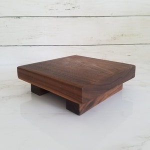 May include: A small, square wooden pedestal with four legs. The pedestal is made of dark brown wood and has a smooth, polished finish.