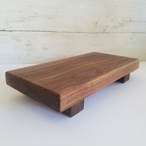 May include: A rectangular wooden serving board with a rich brown finish. The board is elevated on four small, square feet. The wood grain is visible, adding texture to the surface. The board is designed for serving food.