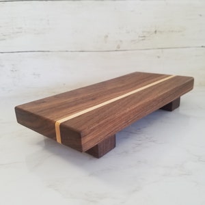 May include: A rectangular wooden serving tray with a light wood inlay. The tray has four small legs and is made of dark wood.