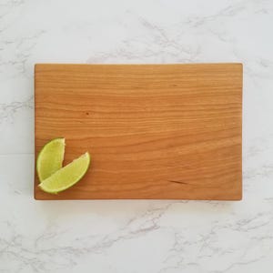 Op de afbeelding: Een klein, rechthoekig houten snijplank van kersenhout. De plank heeft een natuurlijke houtkleur en is afgebeeld met twee limoenpartjes erop.