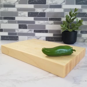 May include: A light-colored rectangular wooden cutting board with a green jalapeño pepper on top. A small potted plant is in the background. The cutting board is made of light-colored wood.