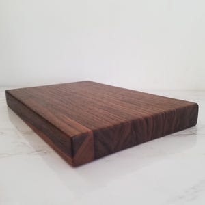 May include: A rectangular wooden cutting board made of dark brown walnut wood. The board has a smooth surface and a slightly raised edge. The board is shown from an angle, with the edge closest to the viewer in focus.
