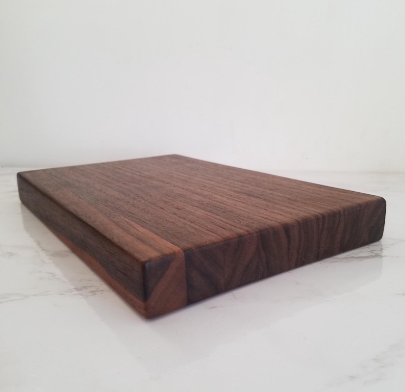 May include: A rectangular wooden cutting board made of dark brown walnut wood. The board has a smooth surface and a slightly raised edge. The board is shown from an angle, with the edge closest to the viewer in focus.
