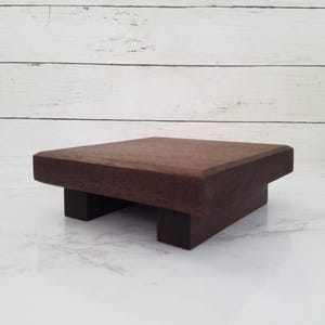 以下が含まれることがあります： 正方形の上面と2つの長方形の脚が付いた、小さな濃い茶色の木製ライザー。ライザーは無垢材で作られており、滑らかで仕上げられた表面を持っています。デザインはシンプルでモダンで、小さなアイテムの表示に適しています。