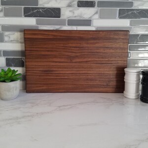 May include: A large, rectangular, brown wooden cutting board with a wood grain pattern. The cutting board is sitting on a white countertop.
