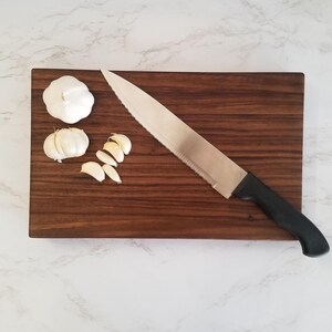 May include: A brown wooden cutting board with a silver knife and three cloves of garlic, one whole and two sliced.