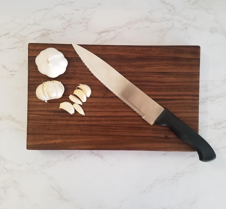 May include: A brown wooden cutting board with a silver knife and three cloves of garlic, one whole and two sliced.