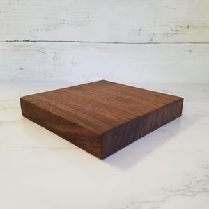 May include: A square wooden block in a dark brown hue, showcasing visible wood grain. The edges are precisely cut. This block could serve as a display stand or a small platform, likely made of hardwood.