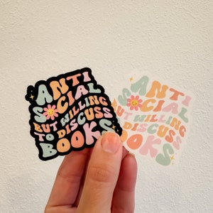 May include: Two stickers with the text "Anti Social But Willing To Discuss Books" in a colorful, retro font. The stickers are in different color schemes, one with a black background and the other with a white background.