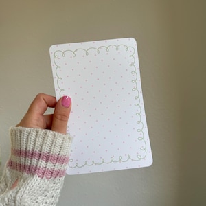 May include: A white rectangular card with rounded corners, adorned with a light green scalloped border and small pink polka dots. The card is held up by a person wearing a white sweater with pink stripes.