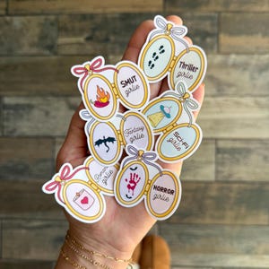 May include: A hand holding a variety of stickers shaped like locket charms. Each charm has a different design and text, including "SMUT girlie", "Fantasy girlie", "Sci-Fi girlie", "Thriller girlie", "Horror girlie", "Romance girlie".
