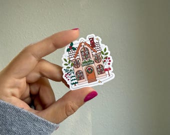 May include: A sticker featuring a whimsical illustration of a bookstore. The building is pink with arched windows, a red door, and a chimney. The windows display books, and the sign reads "BOOKSTORE". The sticker is decorated with festive greenery.