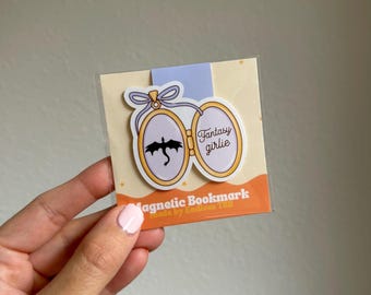 May include: A magnetic bookmark featuring a gold-framed locket design with a black dragon silhouette and the words "Fantasy girlie." The bookmark is attached to a light blue tab and a tan backing with text.