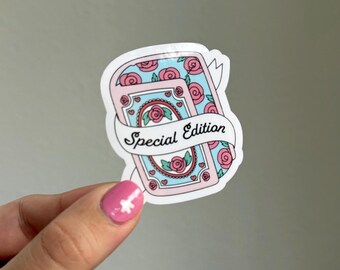 May include: A sticker featuring a design of two playing cards with a light blue background and pink roses. The cards are wrapped with a white banner that reads "Special Edition". The sticker is held by a hand with pink nail polish.