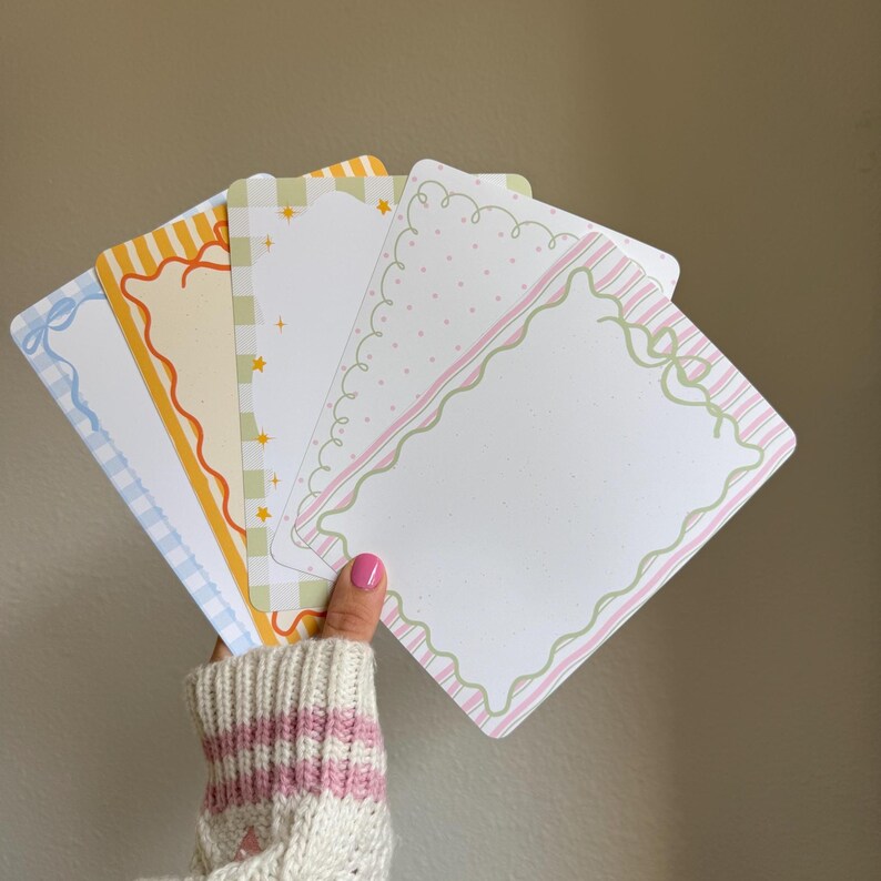 May include: A hand holds a fan of decorative cards with various pastel borders. The cards feature designs like scalloped edges, polka dots, stripes, and bows. Colors include blue, orange, yellow, green, and pink.