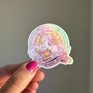 May include: A holographic sticker featuring a unicorn reading a book with a rainbow and stars in the background. The text reads "Magical Reader's Society".