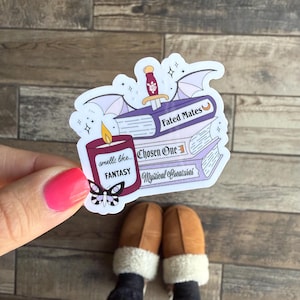 May include: A sticker featuring a stack of books with titles "Fated Mates", "Chosen One", and "Mystical Creatures". A purple candle with the text "smells like... FANTASY" is in front of the books. A bat with a dagger is above the books.